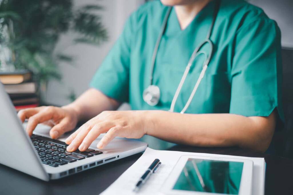 a medical professional in mint green scrubs with a stethoscope around their neck types on a laptop with pen and paper beside it on the table