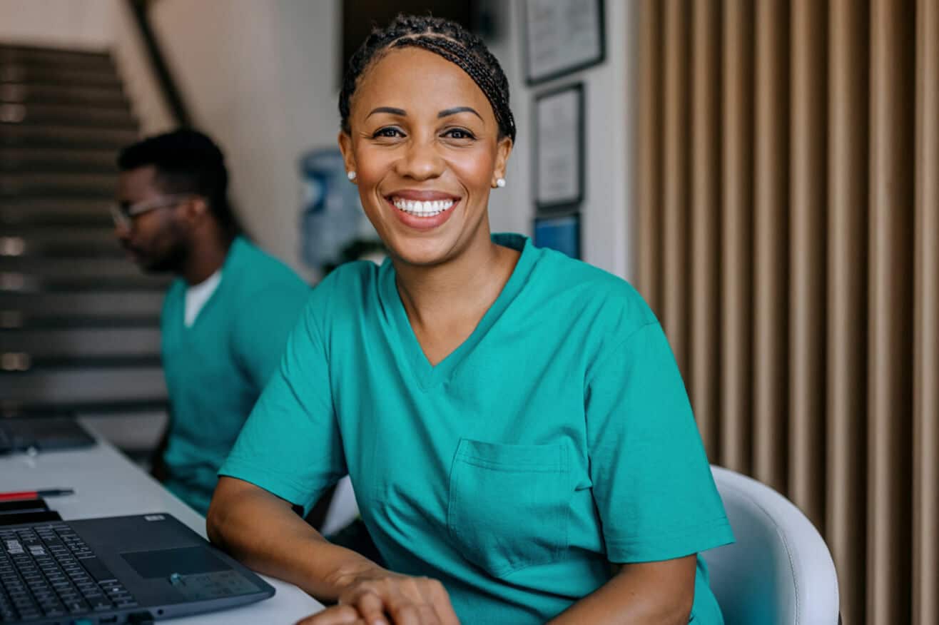A smiling woman in green scrubs sits at a desk, working on a laptop, exuding professionalism and warmth.