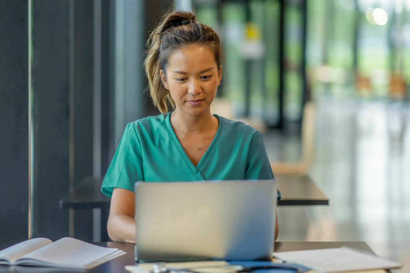 A female nurse in scrubs is seated at a table, working on a laptop, focused and engaged in her tasks.