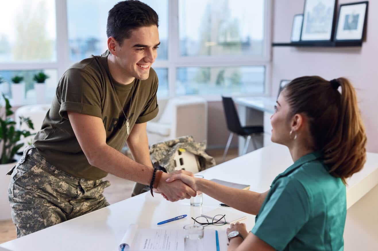 military service member with short dark hair and wearing a brown tshirt smiles and shakes hands across a desk with a female healthcare worker wearing blue scrubs