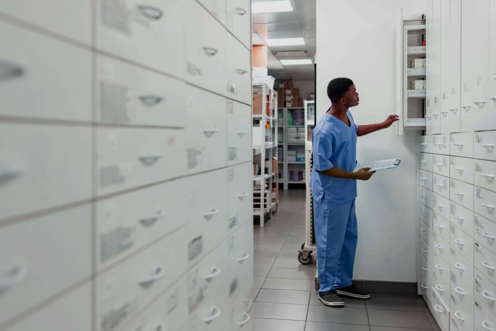 male pharmacy tech in blue scrubs holds a clipboard and searches a large wall of cabinets for medicine in the backroom of a pharmacy