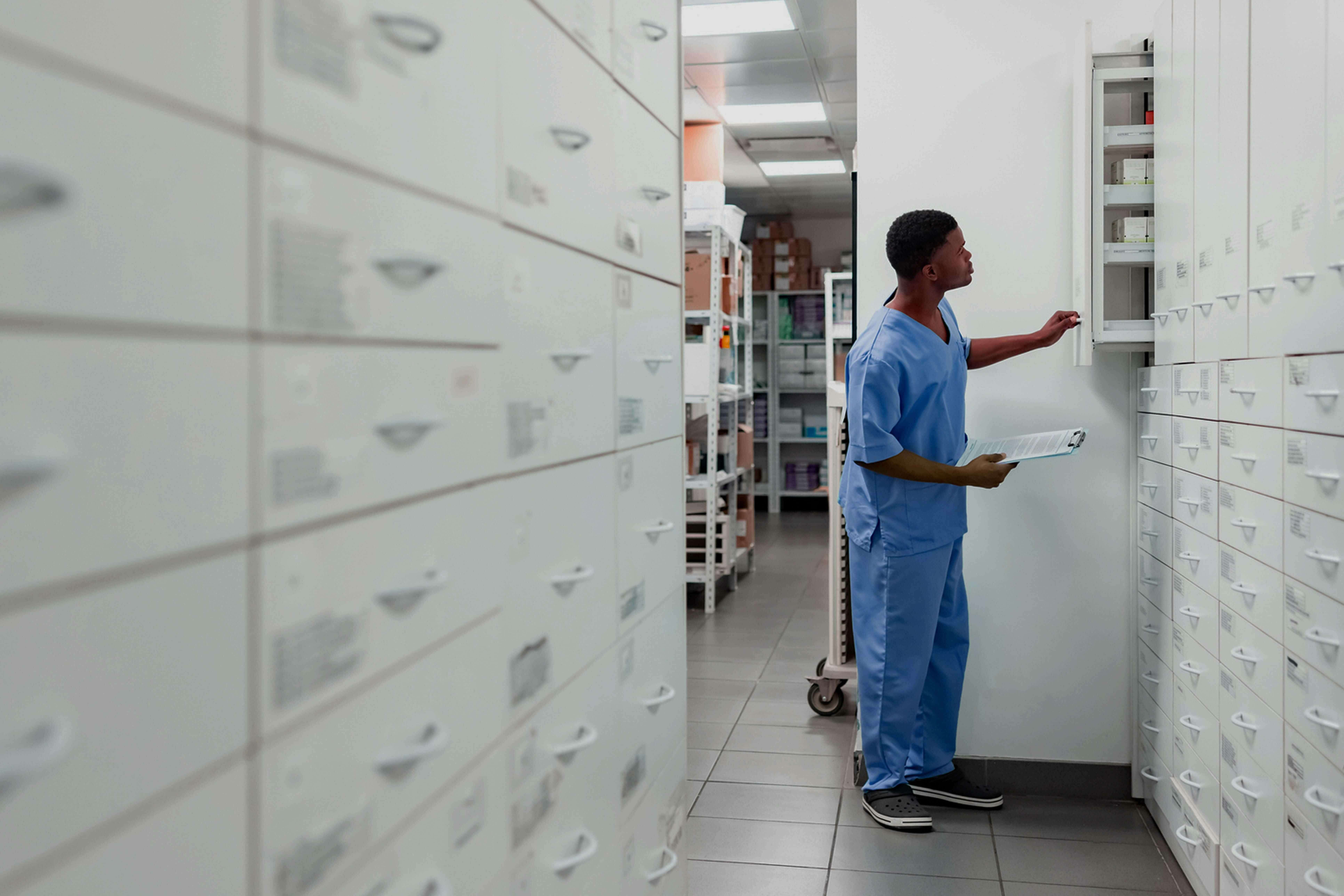 male pharmacy tech in blue scrubs holds a clipboard and searches a large wall of cabinets for medicine in the backroom of a pharmacy