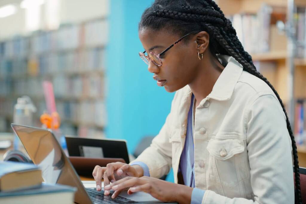 a student with long dark braided hair and glasses works on a laptop in a library wearing a white jean jacket
