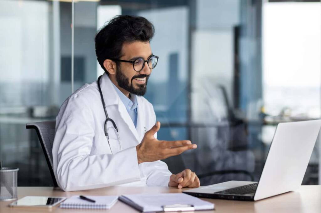 man in a white doctors coat with dark hair and beard wearing glasses consults with a patient via laptop