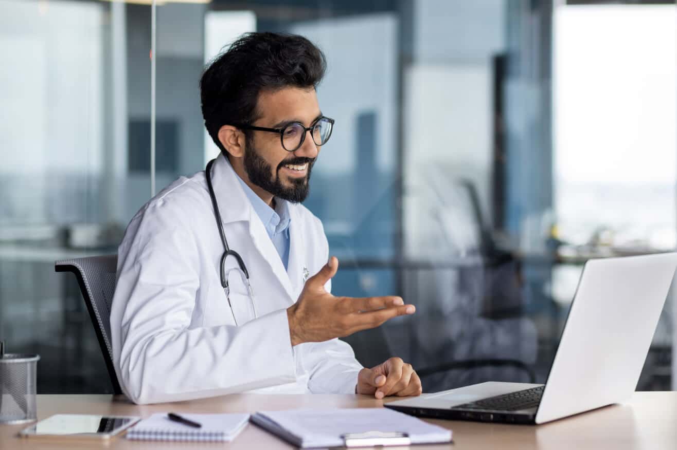 man in a white doctors coat with dark hair and beard wearing glasses consults with a patient via laptop