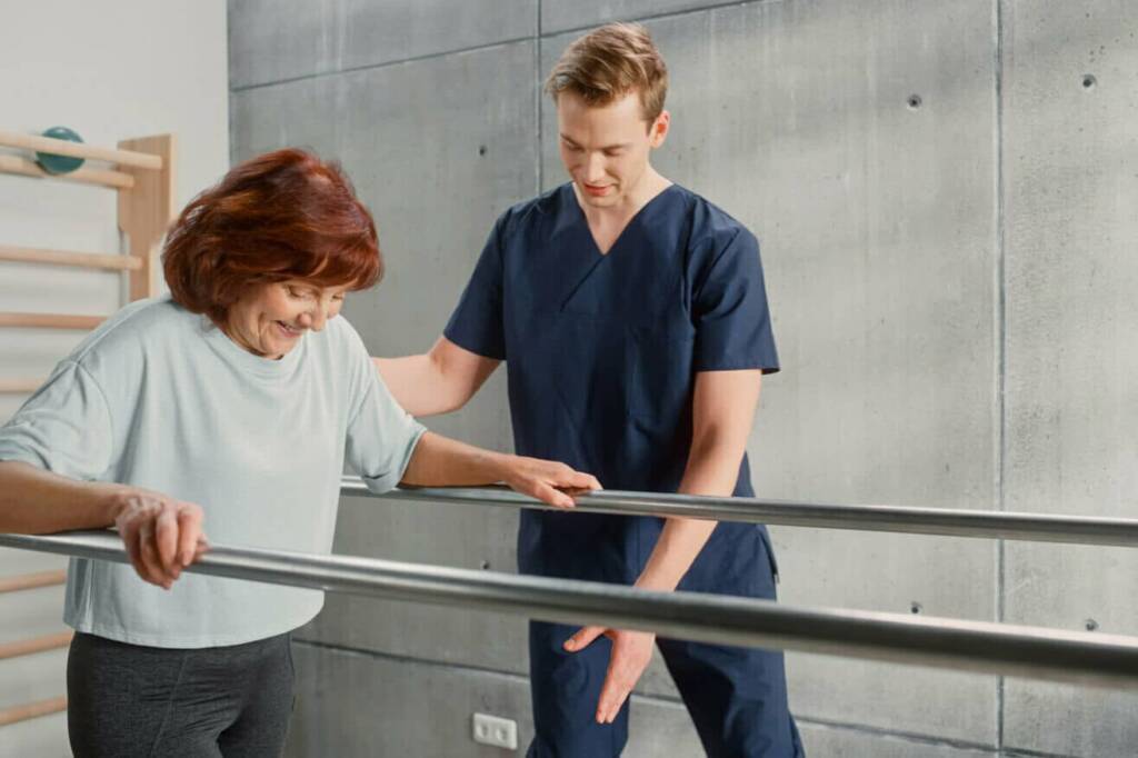 a male physical therapists assistant in dark blue scrubs with short blonde hair helps a red-headed female patient walk as she holds onto metal bars