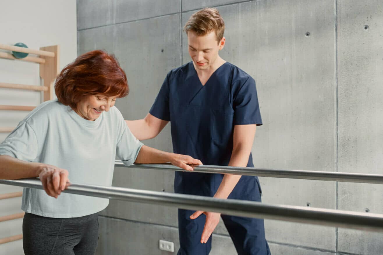 a male physical therapists assistant in dark blue scrubs with short blonde hair helps a red-headed female patient walk as she holds onto metal bars