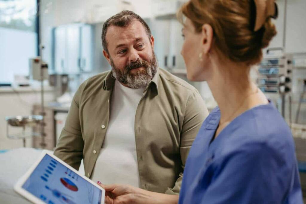 a female doctor in blue scrubs holds a tablet with data on it and speaks to a male patient with a khaki buttondown shirt