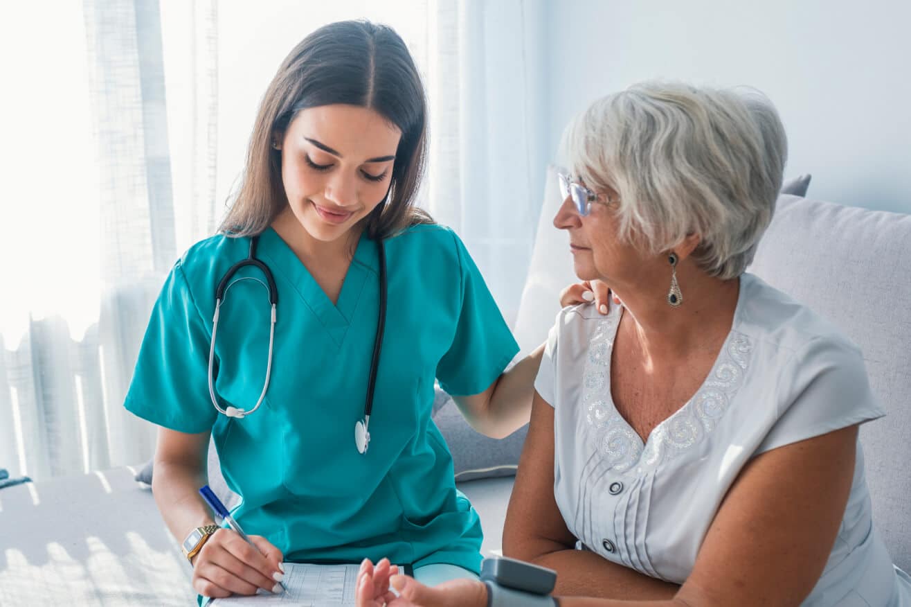nurse with long brown hair writes on paperwork while tending to an older female patient with short grey hair