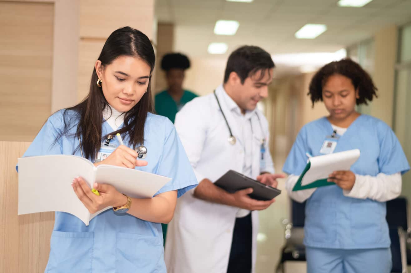 a pre-med student stands in the foreground wearing blue scrubs and takes notes on a packet of papers while a doctor and second student stand behind her reviewing more paperwork