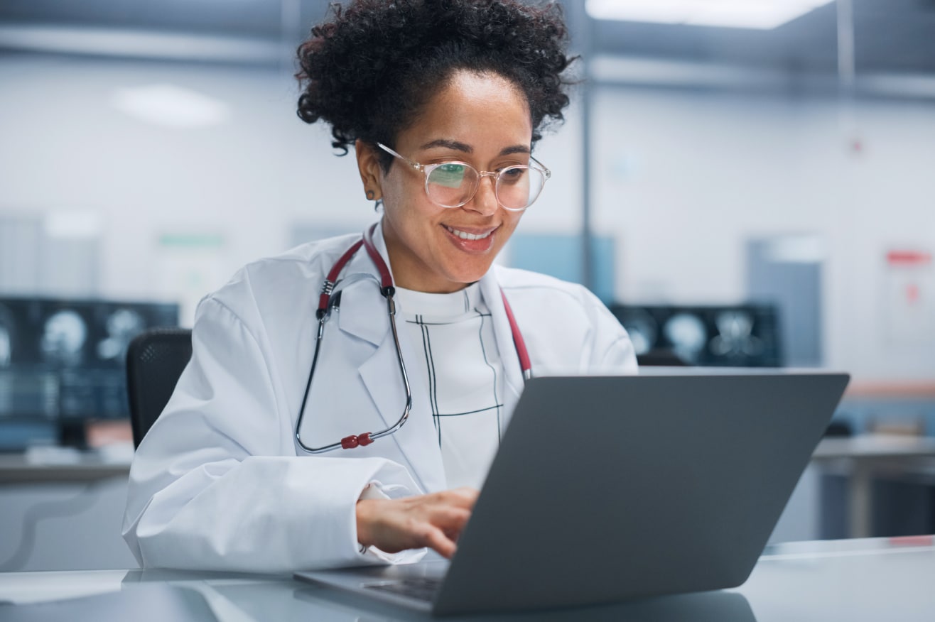 doctor with dark hair and glasses wears a white lab coat and smiles as they work on a laptop in a lab setting