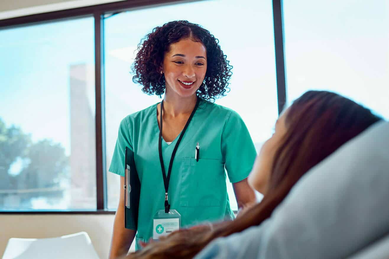 nurse with curly black hair stands by a patient lying in bed, smiling at the patient