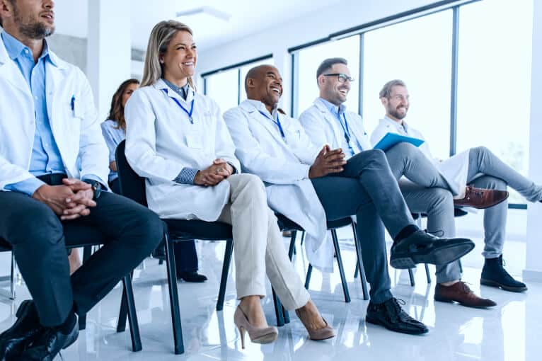 a group of people wearing white lab coats sit in a row of chairs as an audience looking at something outside of the image, they're all smiling and laughing