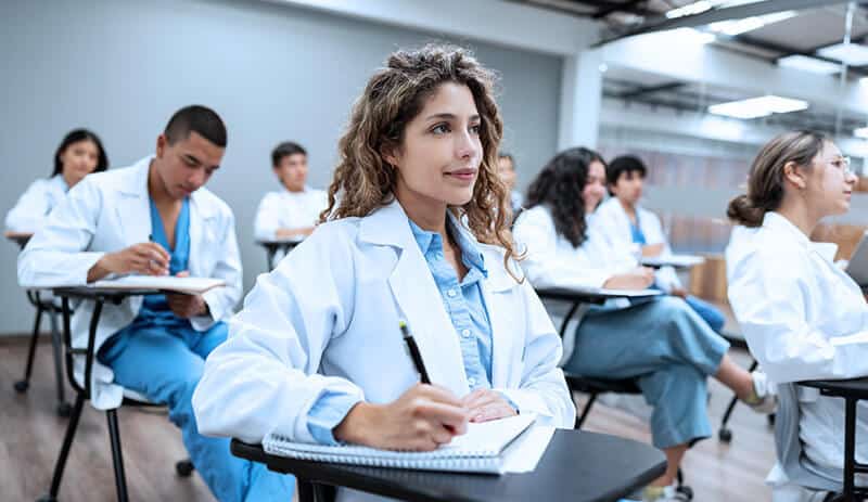 a medical student with long curly hair sits in a classroom at a small desk taking notes on a pad of paper