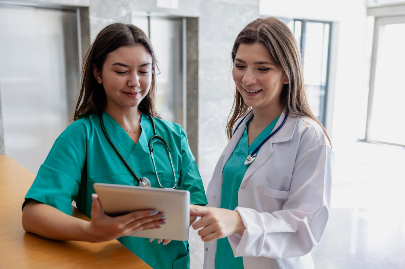 two female healthcare professionals with long straight hair stand together in a medical building hallway reviewing a tablet together