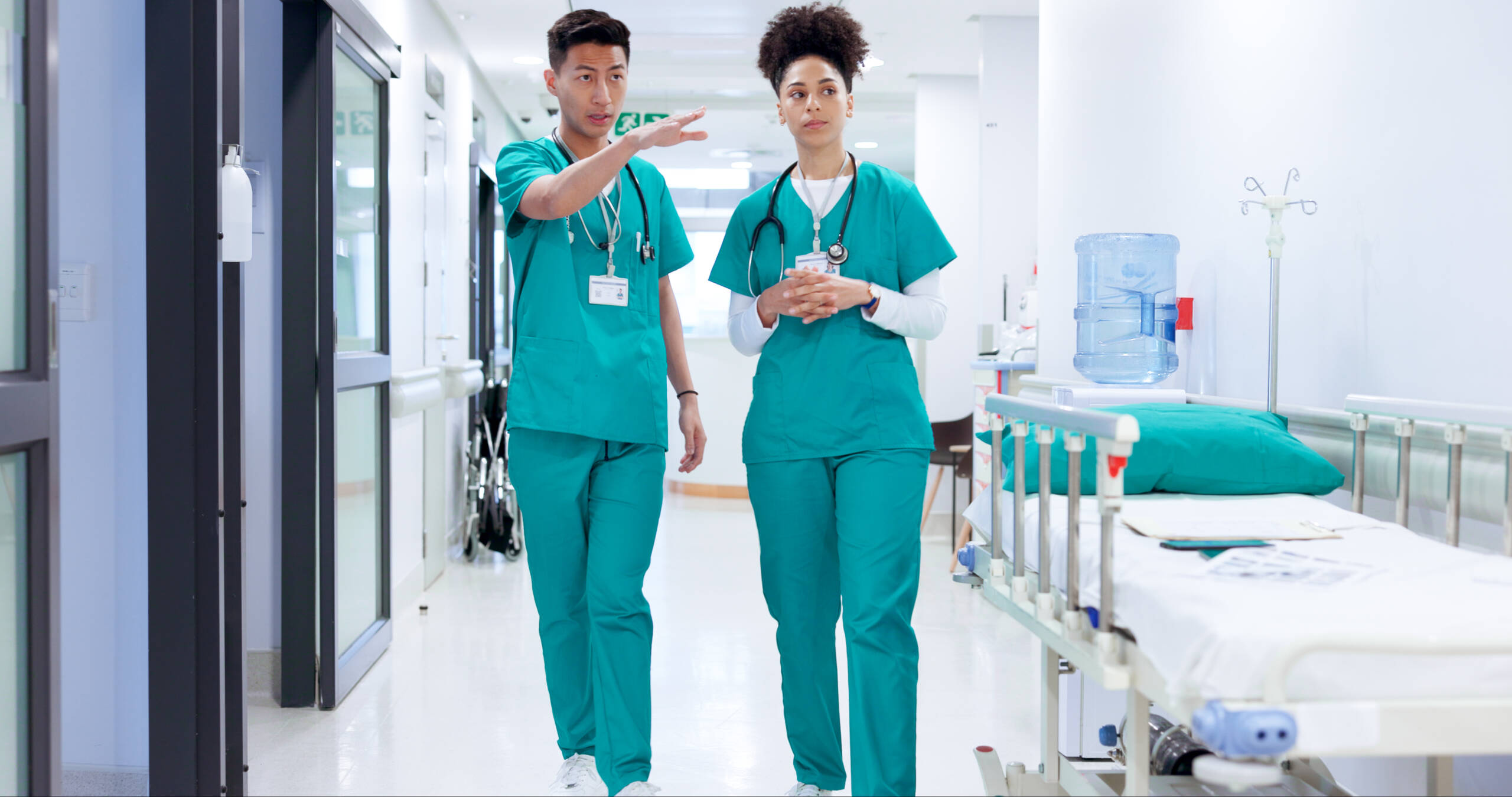 Two nurses walking side by side down a hospital hallway, engaged in conversation.