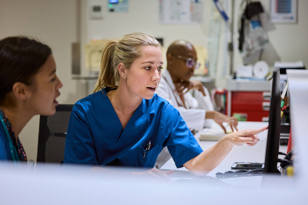 A nurse points at a computer screen, indicating information or data displayed on it.