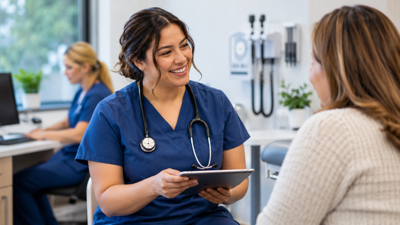 Medical assistant in blue scrubs speaking with a patient in a clinic during Medical Assistants Week 2026.